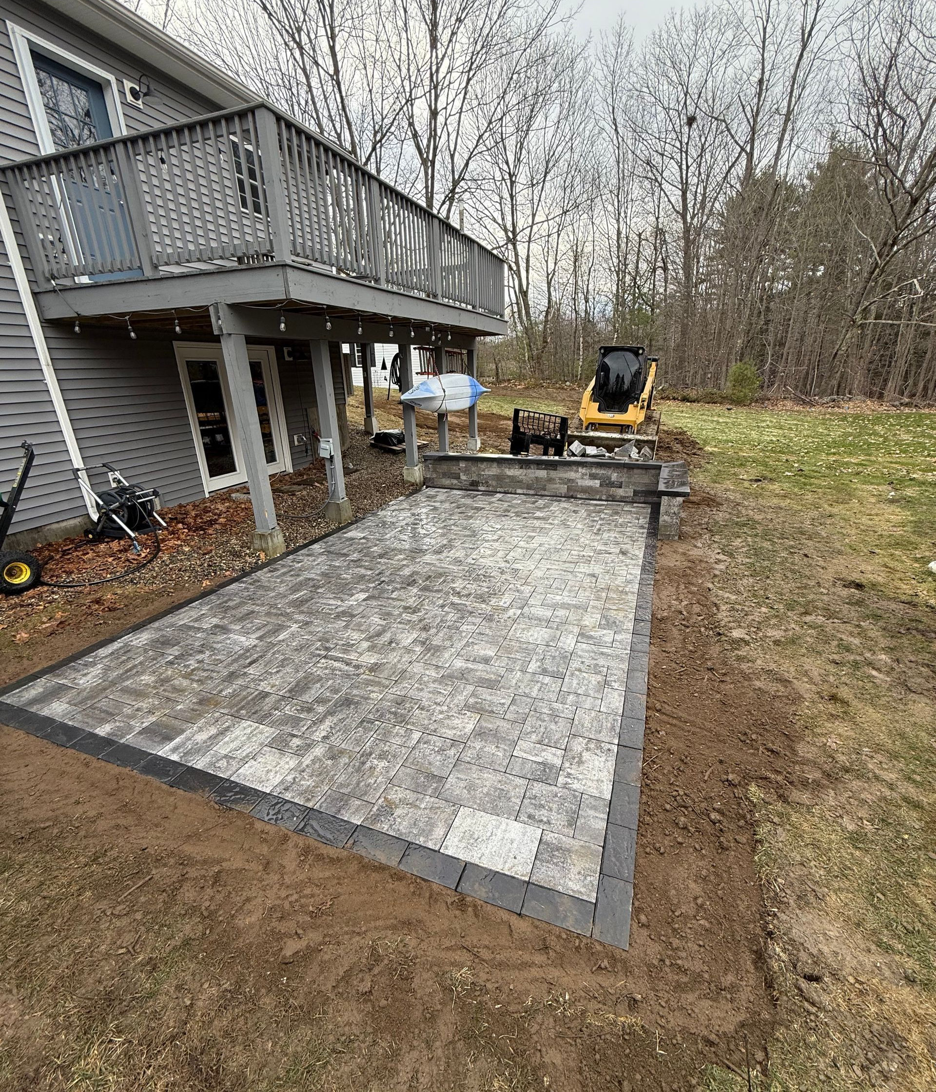 Backyard paver patio beside a gray house with a deck and a compact excavator nearby. 