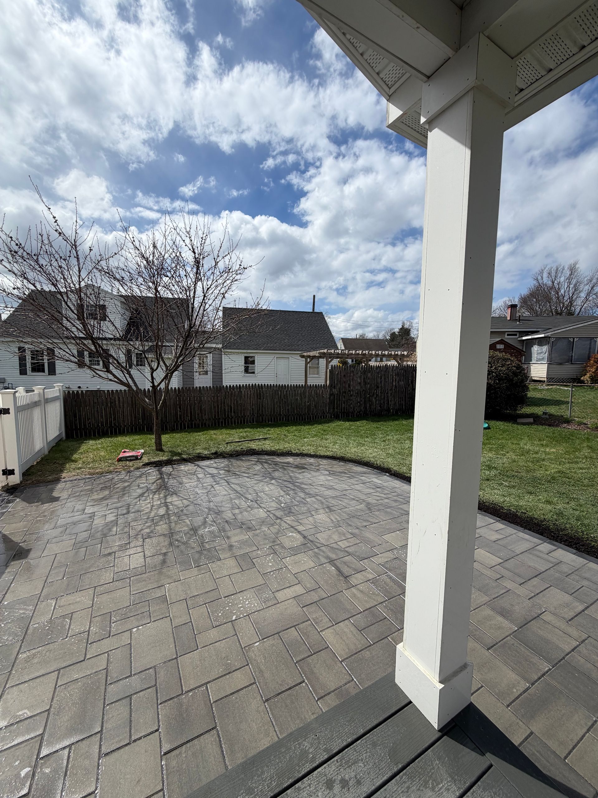 Covered backyard patio with gray pavers, lawn, fence, and cloudy sky. 