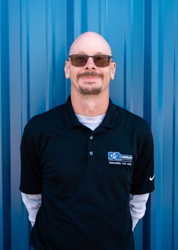 A bald man wearing sunglasses and a black shirt is standing in front of a blue wall.