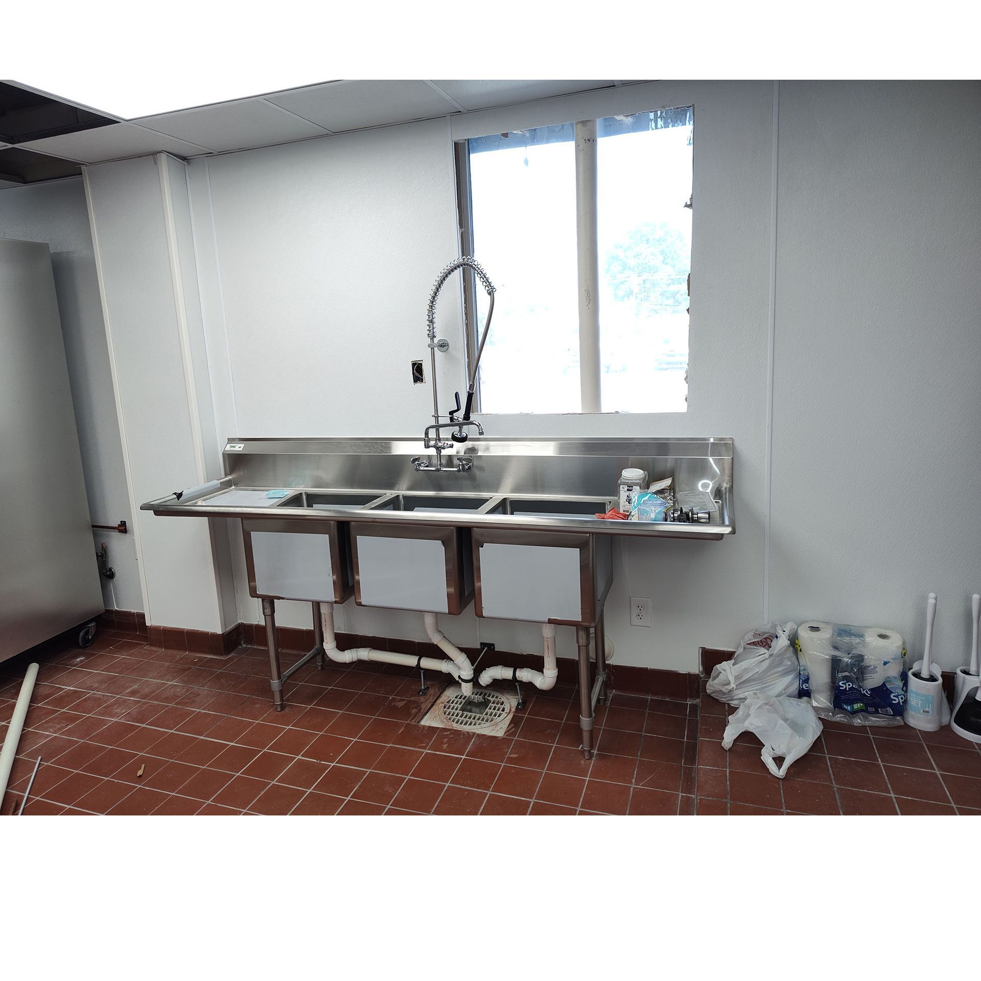 Stainless steel triple sink in a commercial kitchen, beneath a window. Red tile floor and white walls.