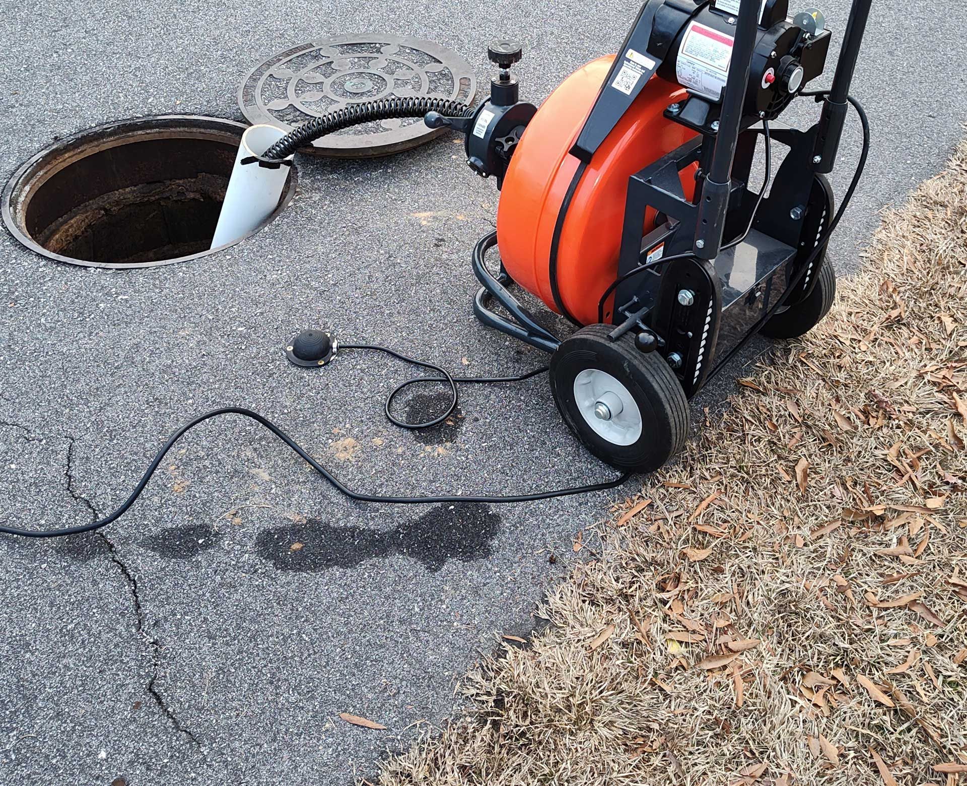 Sewer line cleaning equipment next to an open manhole on asphalt. Orange reel, black cable, grey lid.