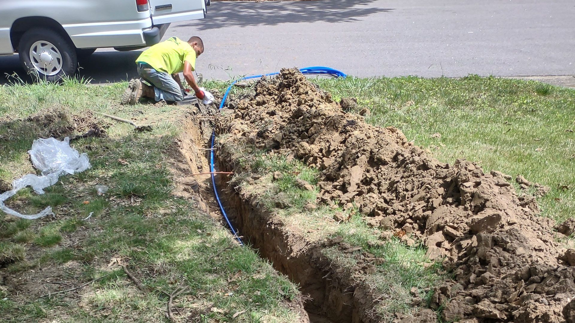 Man kneeling, installing blue pipe in trench on grassy lawn next to a street. Soil piled nearby, truck in background.