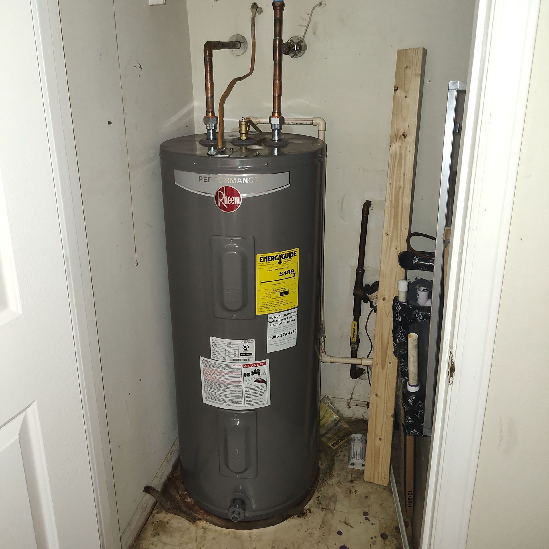 A gray Rheem water heater in a utility closet with pipes and a wooden plank.