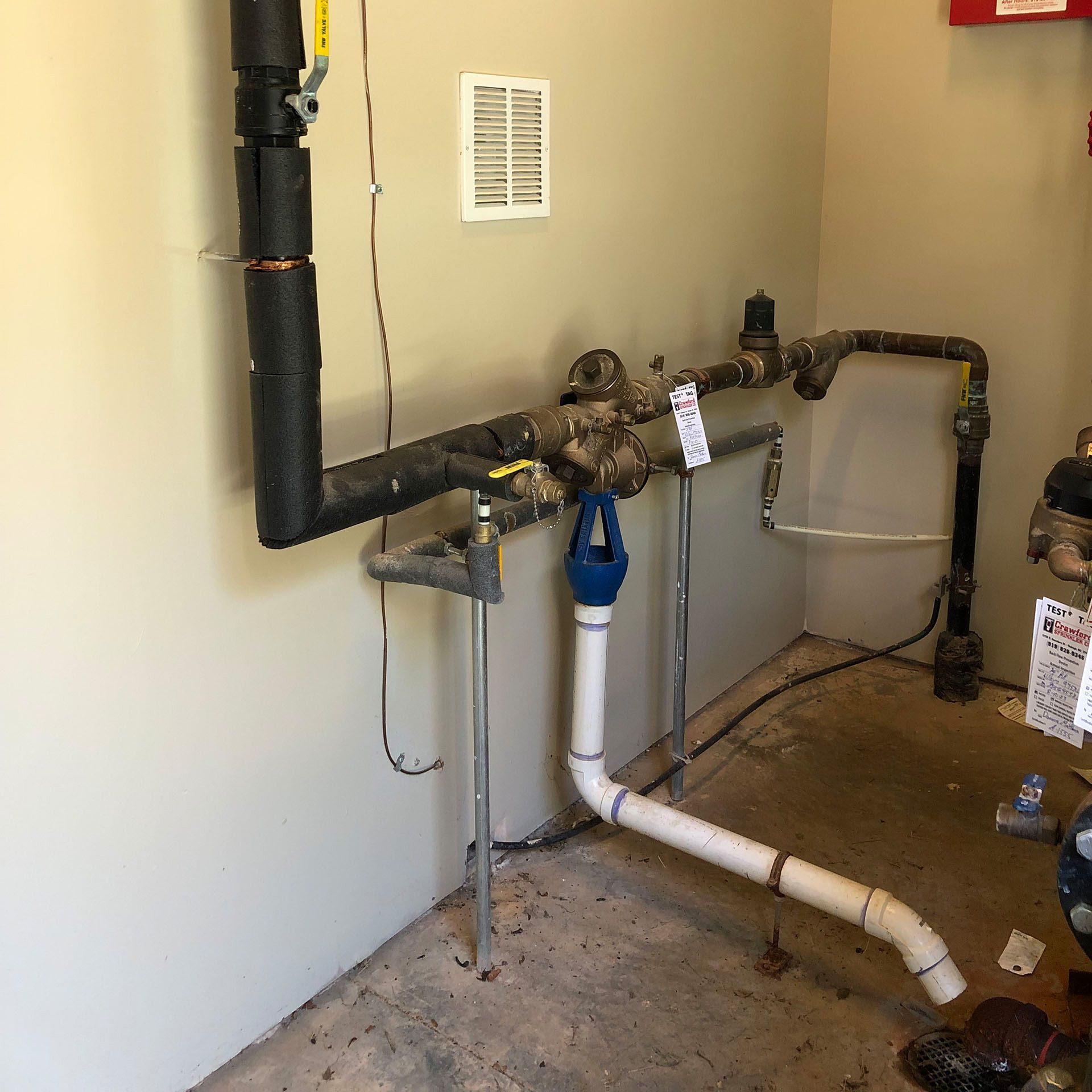Piping system in a beige-walled utility room, including insulated black pipes, valves, and a white PVC drain pipe.