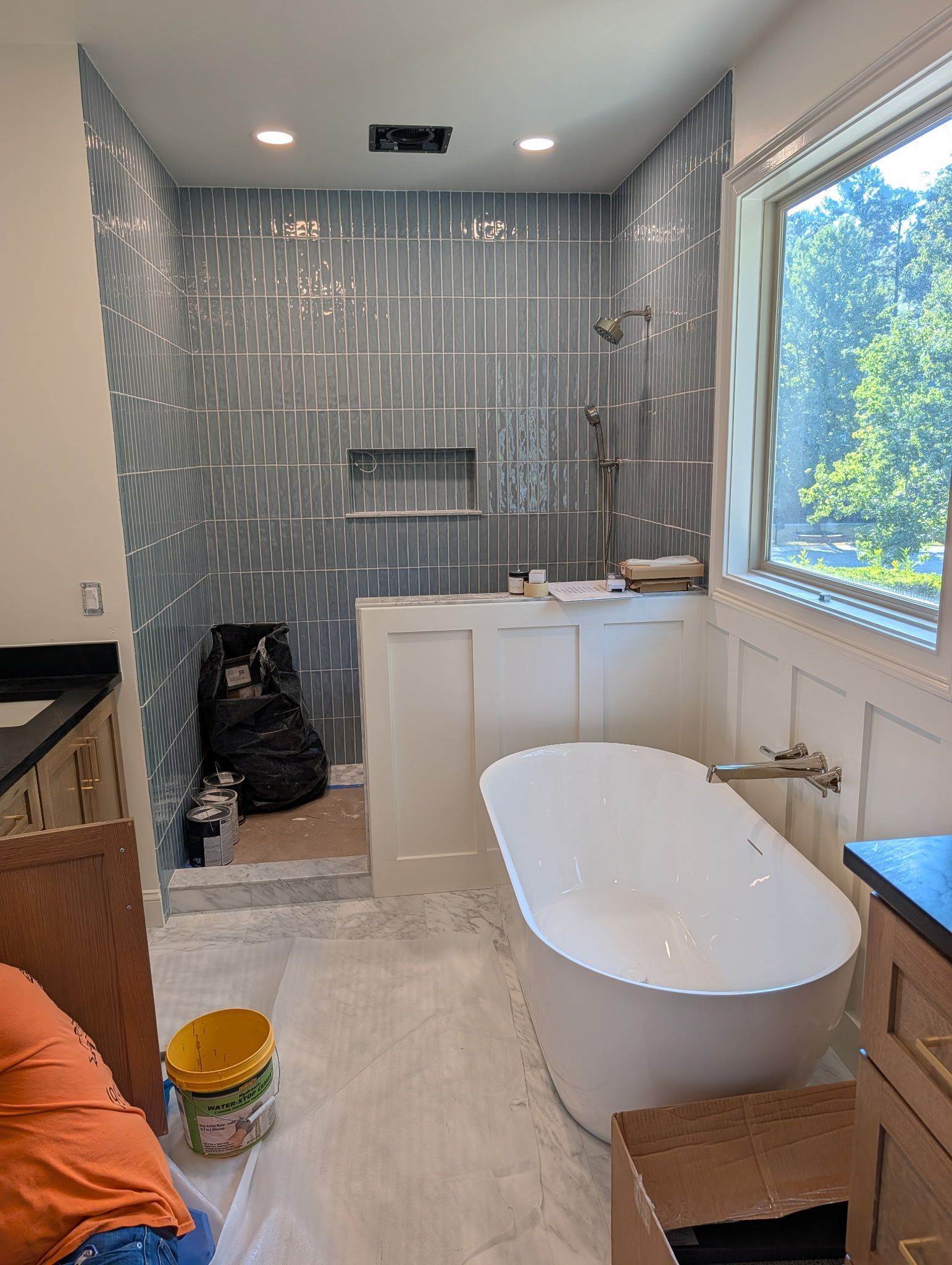 Modern bathroom with freestanding tub, large window, gray tile shower, and renovation materials on the floor