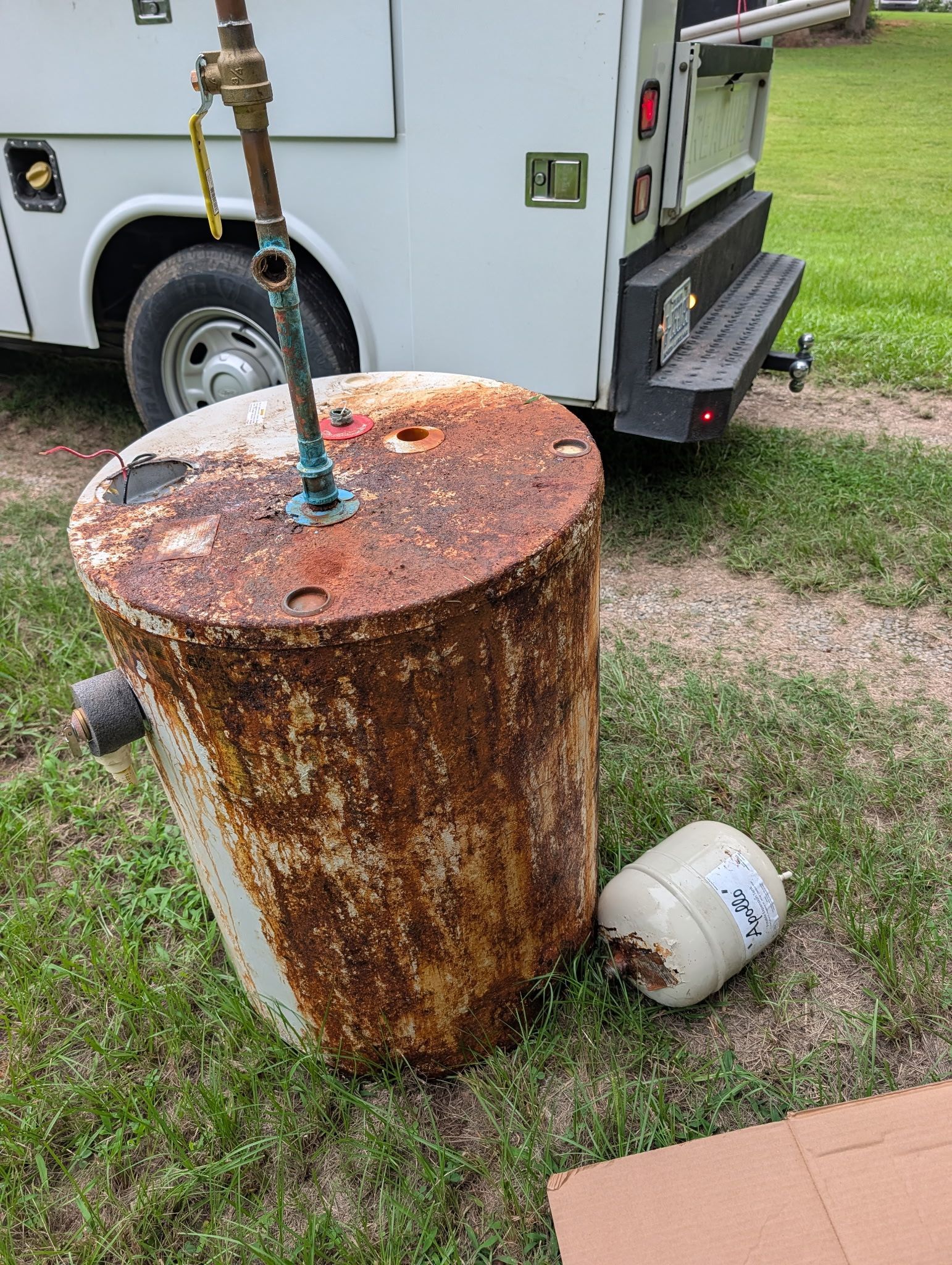 Rusty metal drum with a hose on top, beside a truck on grass; a white bucket леж beside it.
