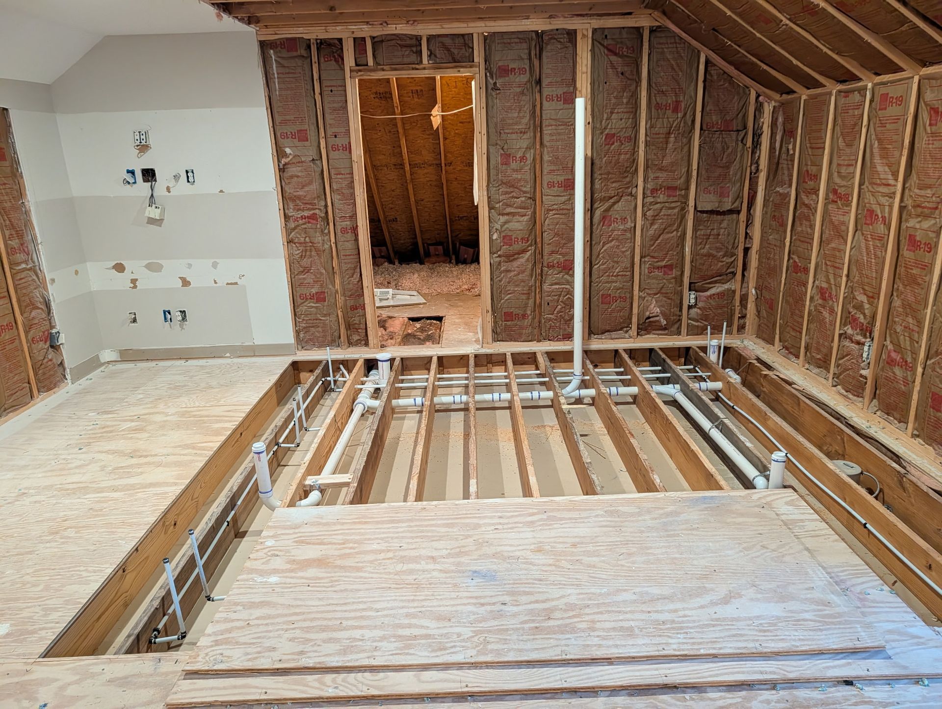 An unfinished attic room showing floor joists, exposed insulation in the walls, and visible white plumbing pipes.