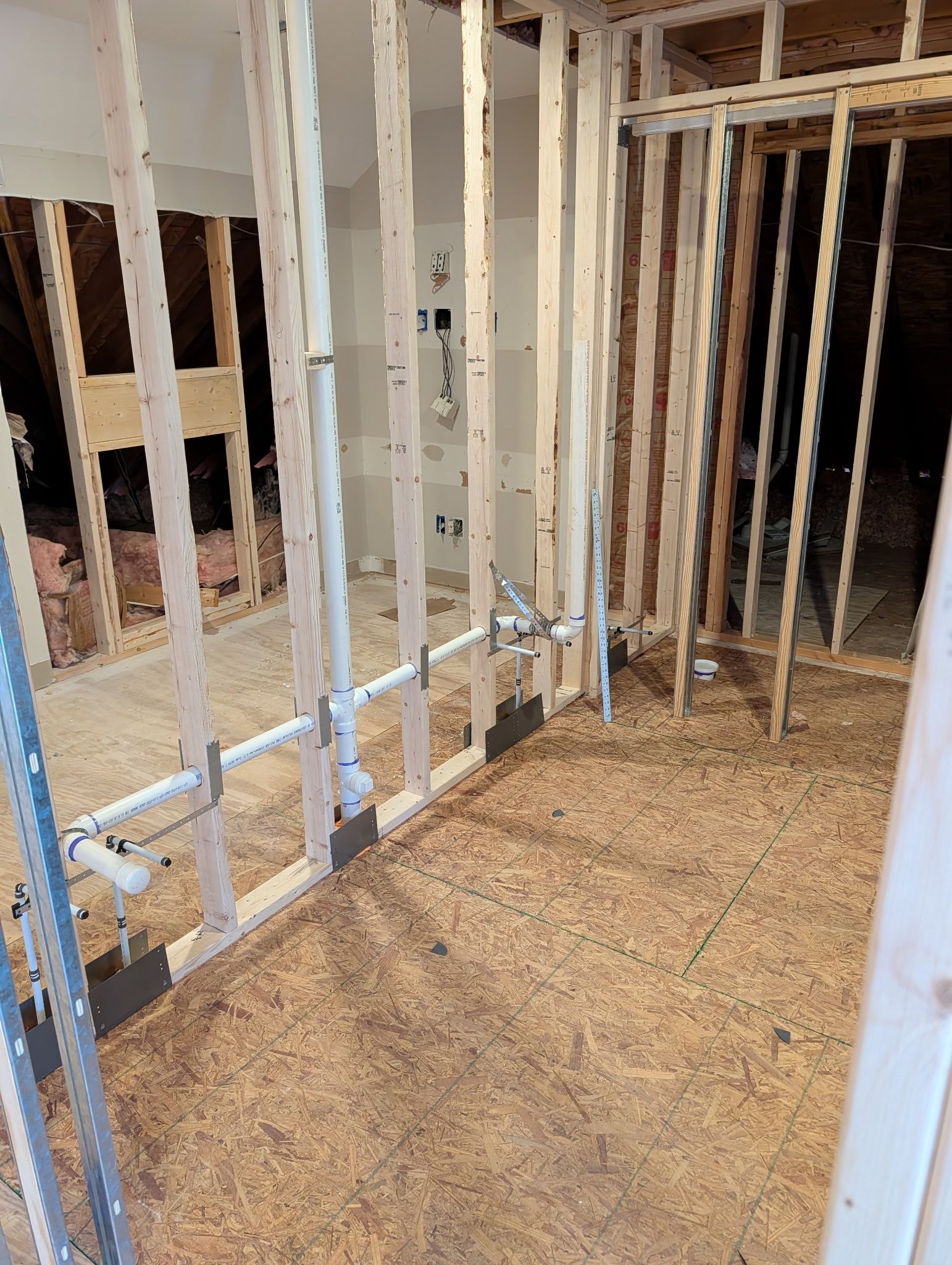 An interior room under construction with exposed wooden wall studs and plumbing pipes installed along the floor.
