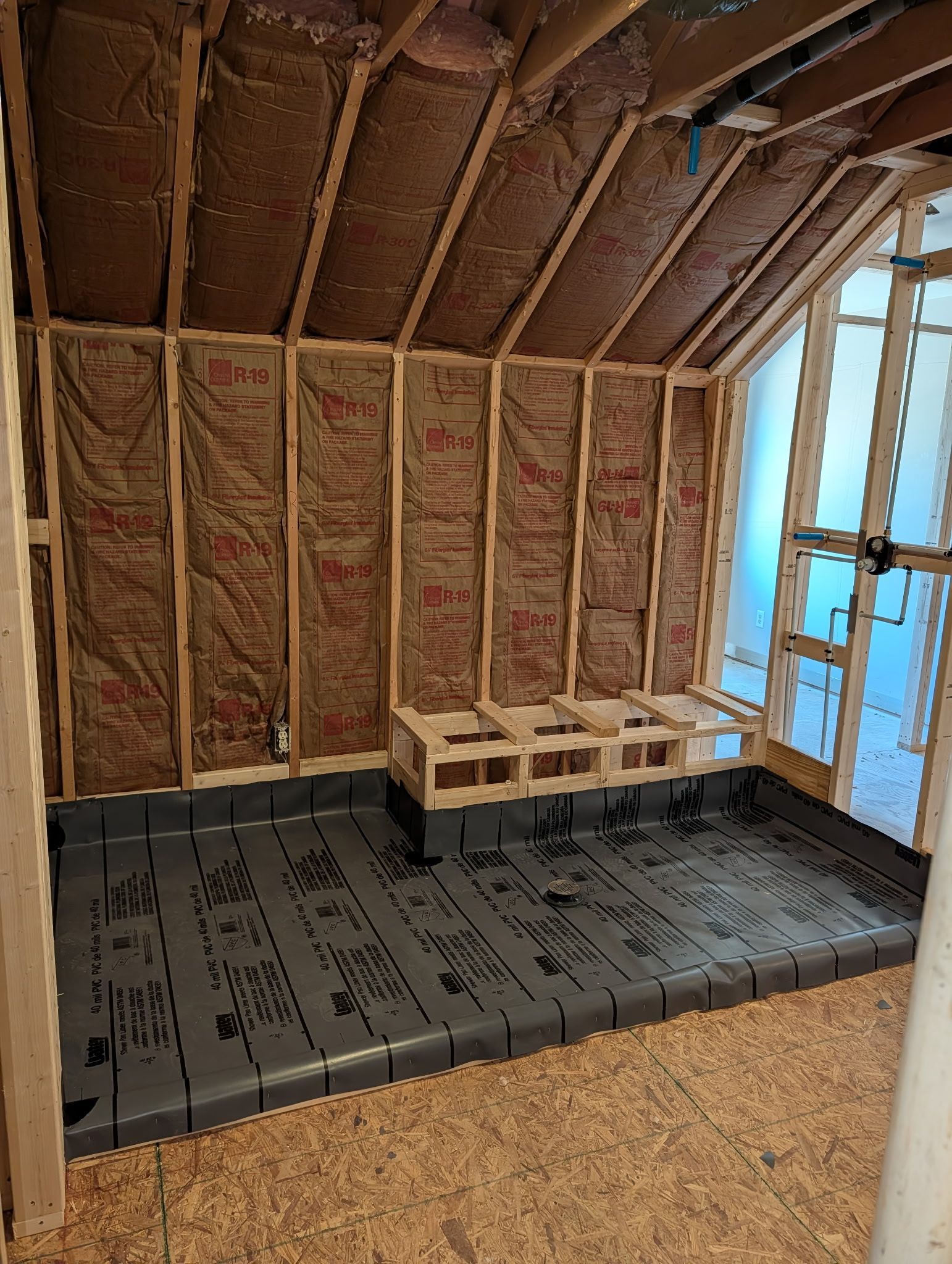 Unfinished room interior featuring wood framing, insulation, a dark gray shower pan, and a built-in wooden shower bench.