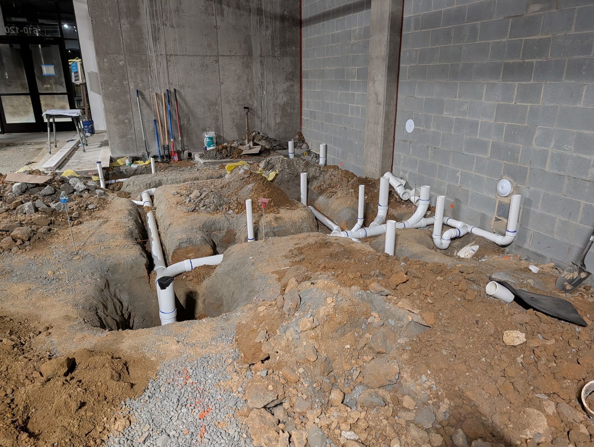 A construction site showing white PVC plumbing pipes installed in deep trenches inside an unfinished room.
