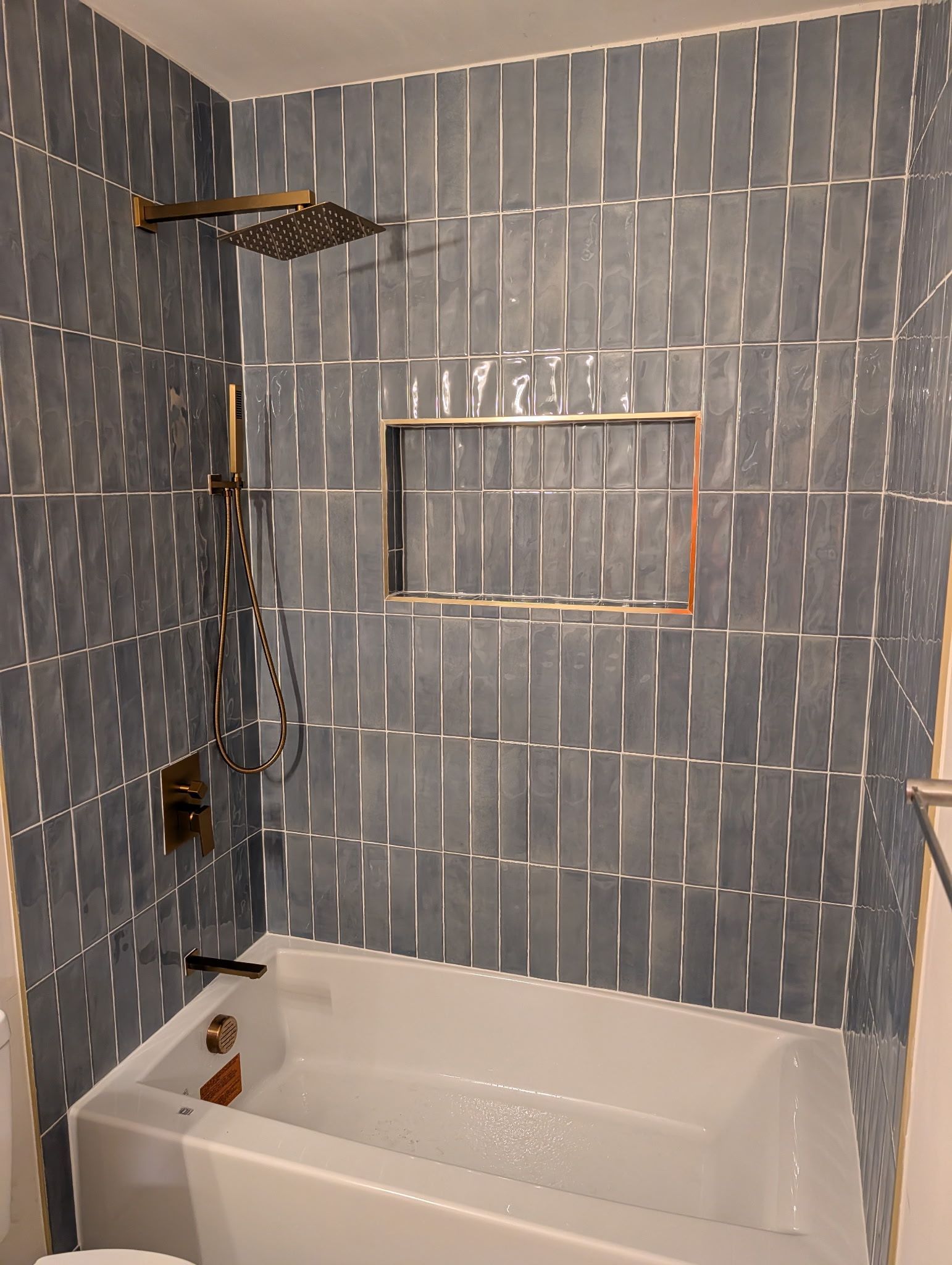 Shower area featuring dark blue vertical tiles, a built-in shelf, and brass shower fixtures above a white bathtub.