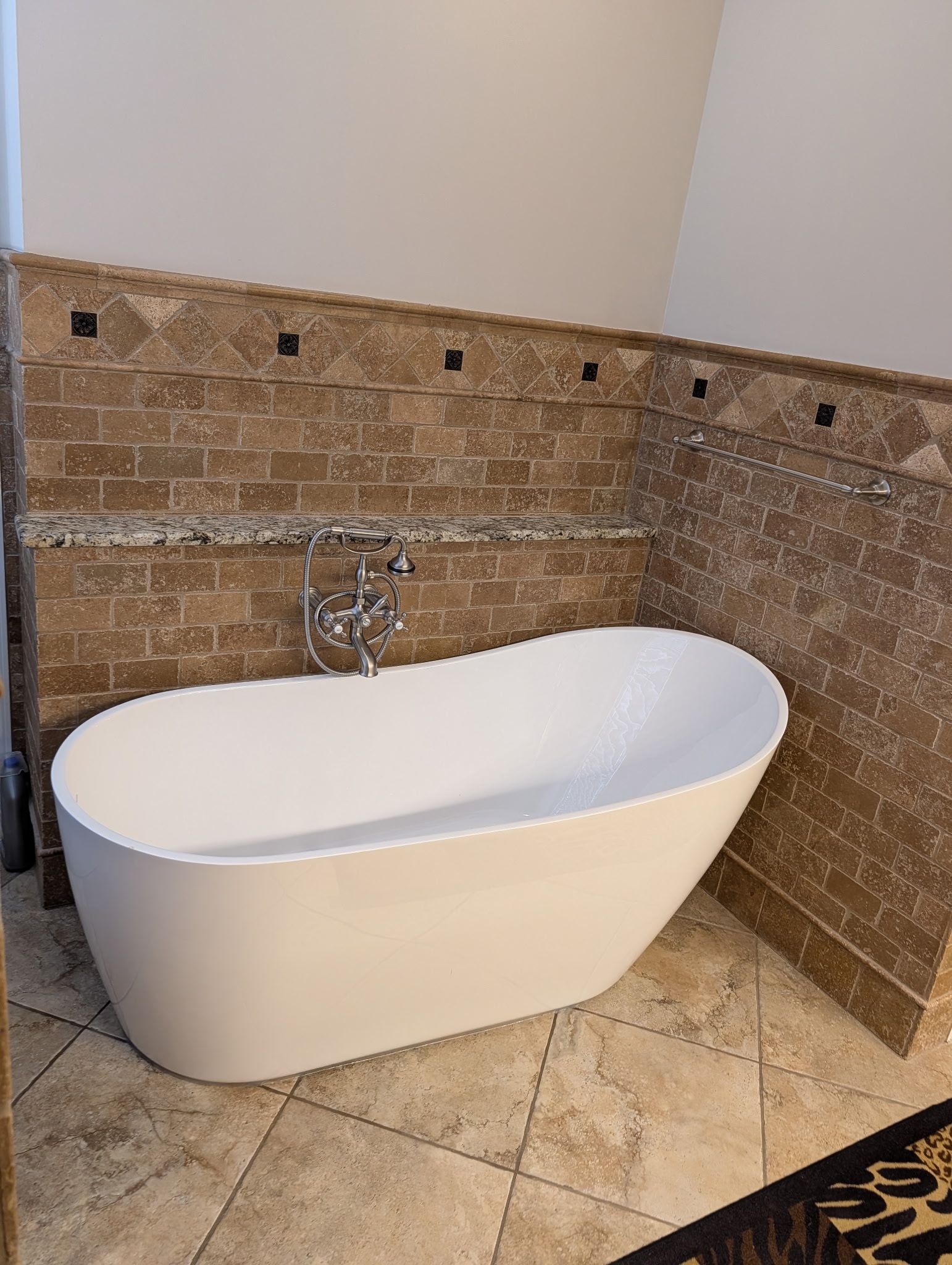 A white freestanding bathtub sits against a corner tiled with textured brown brick and decorative stone accents.