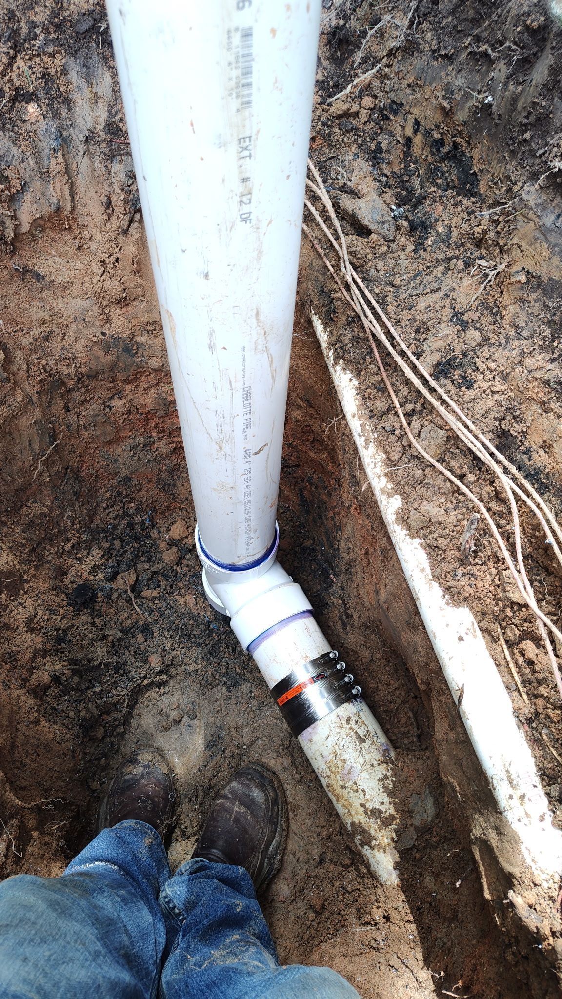A white PVC pipe connected to a trench drain in a dirt excavation, viewed from above by someone wearing boots.