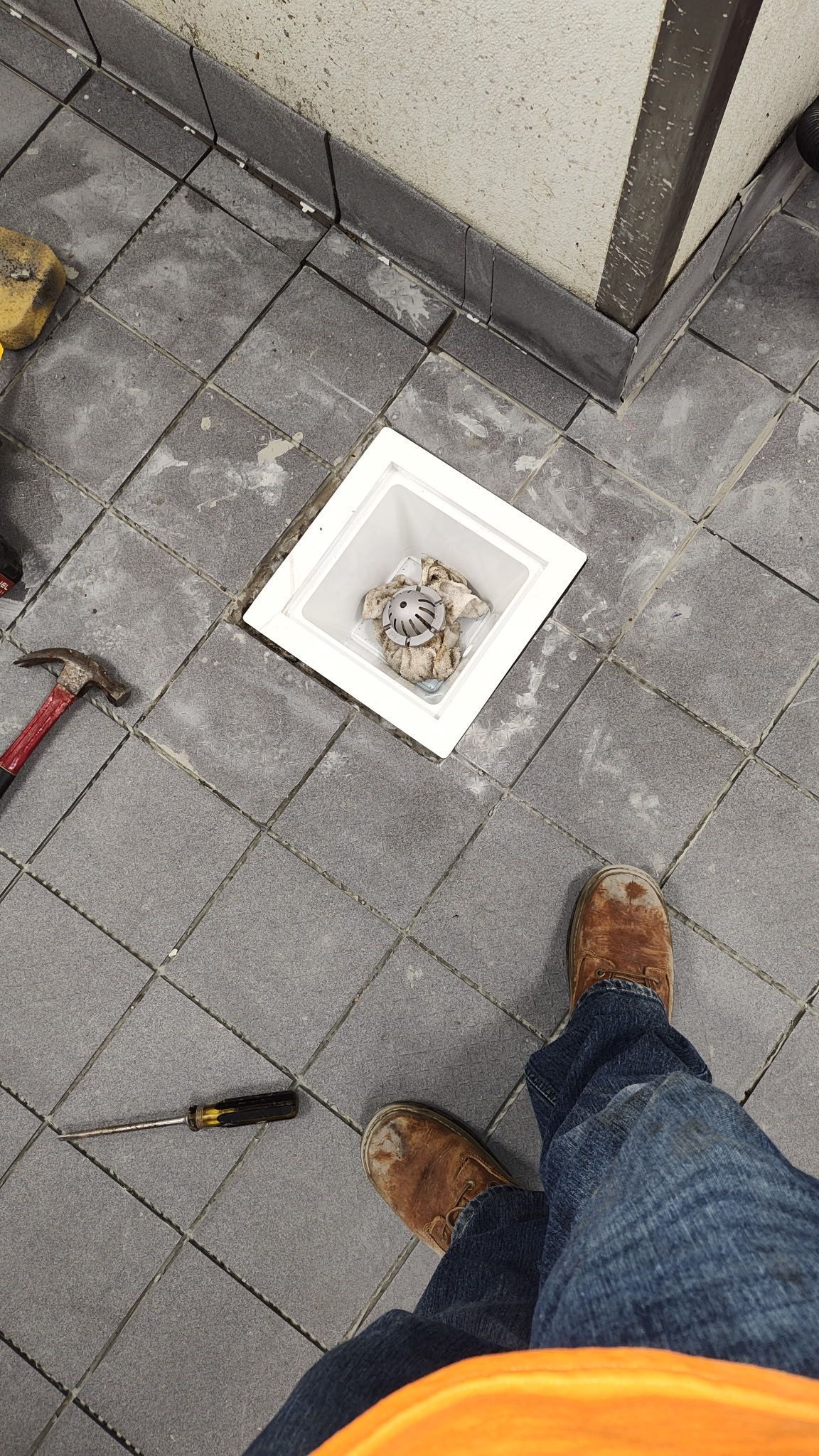 A square plastic drain box set into grey paver stones, with a hammer and screwdriver nearby on the patio.