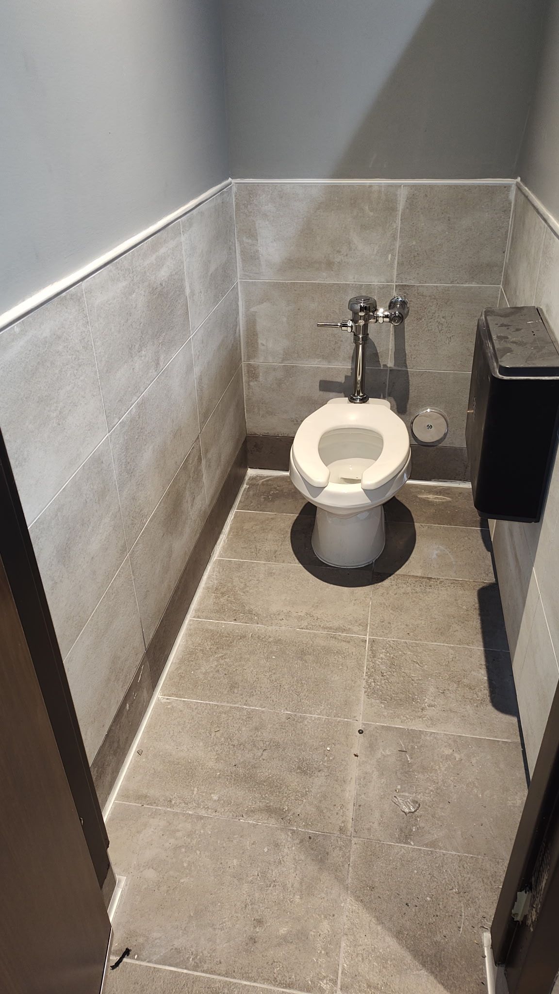 A floor-mounted toilet inside a small stall with light-colored tile walls and gray stone-look flooring.