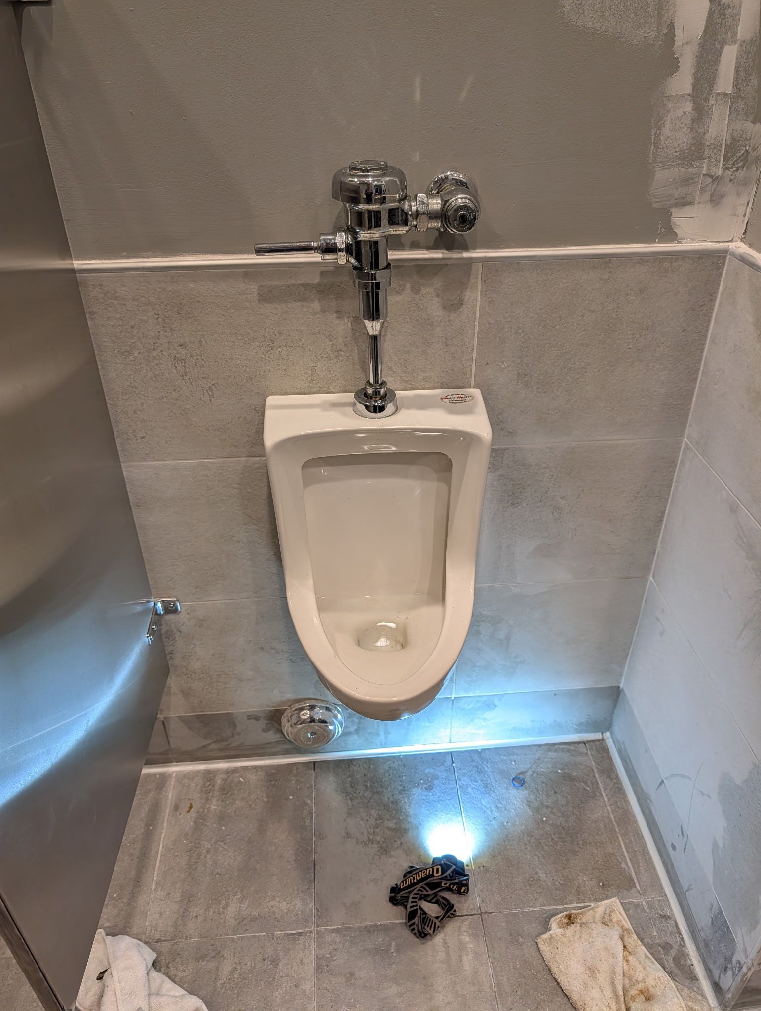 A wall-mounted white urinal in a restroom with a headlamp on the floor shining a bright light toward the fixture.
