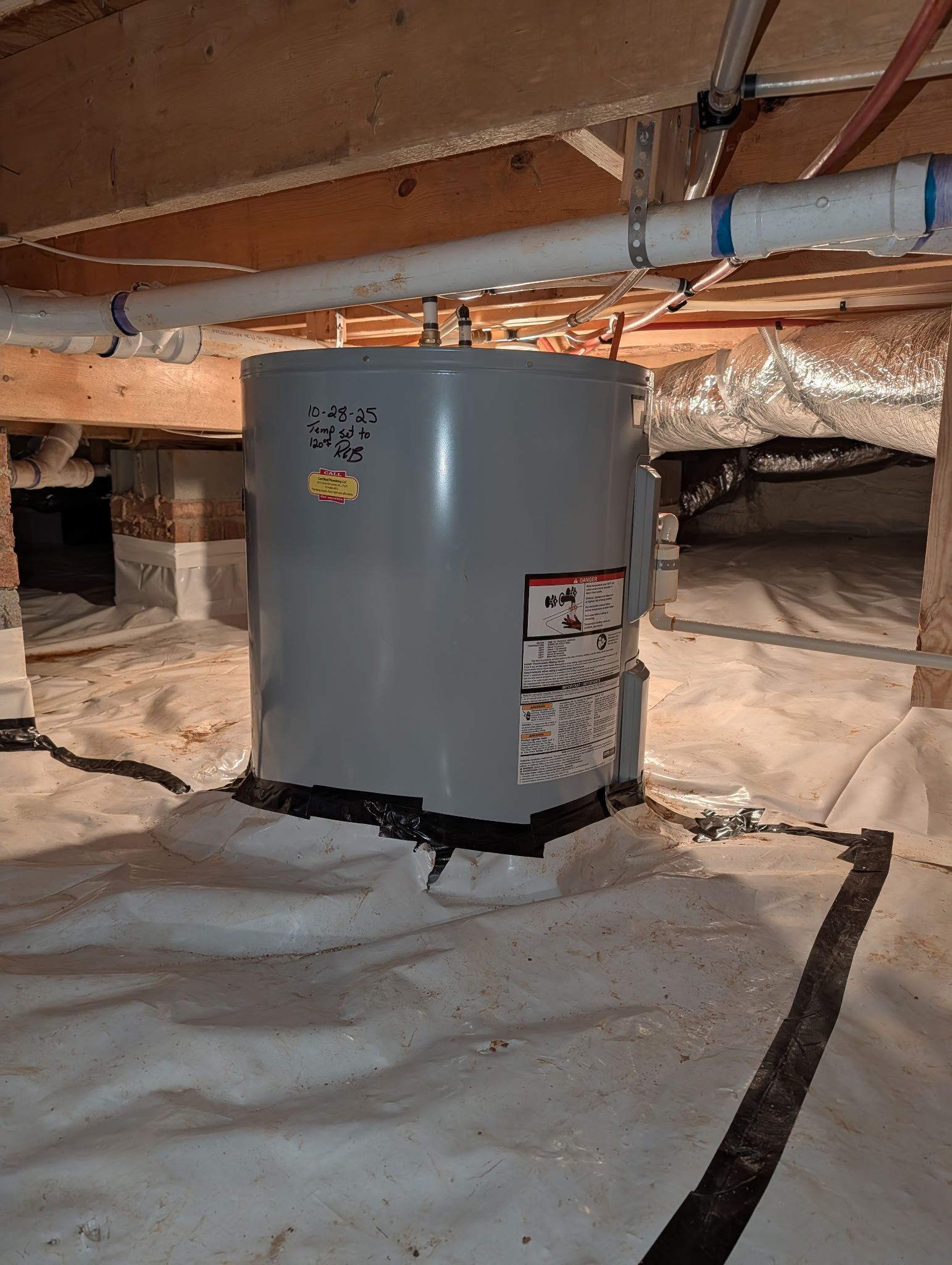 A grey water heater sits on a plastic vapor barrier in a dimly lit crawlspace beneath exposed wooden floor joists.