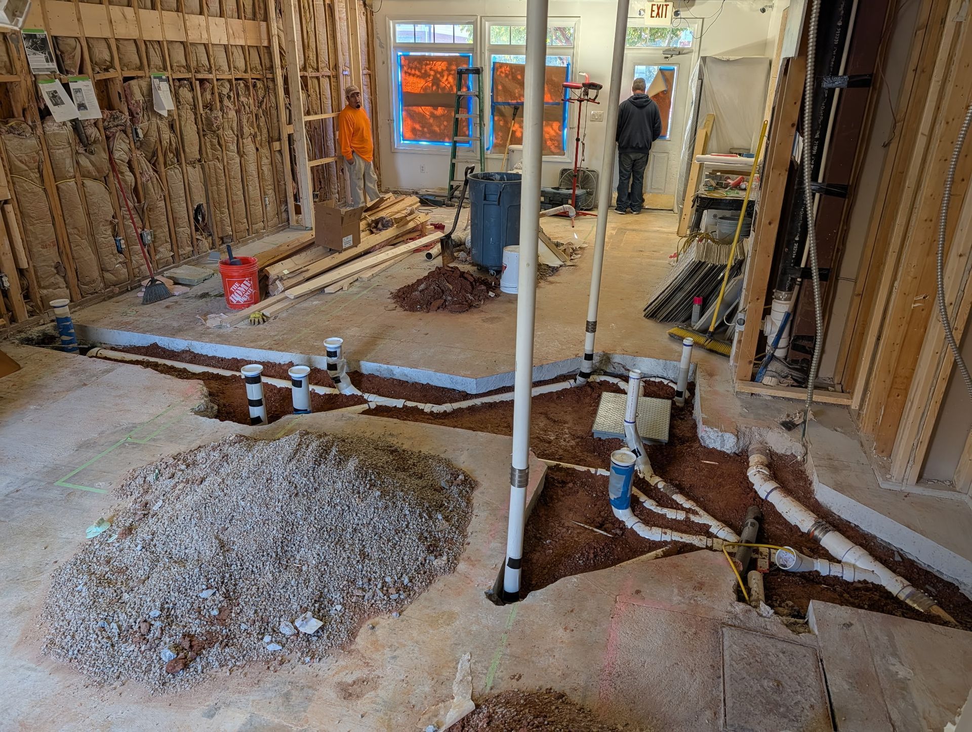 Interior construction site with plumbing pipes laid in trenches, a gravel pile, and exposed wall framing.