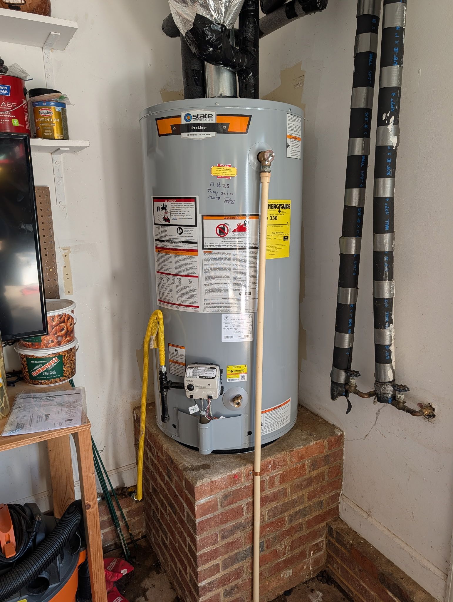 A grey gas water heater stands on a brick pedestal in a garage next to insulated vertical pipes.
