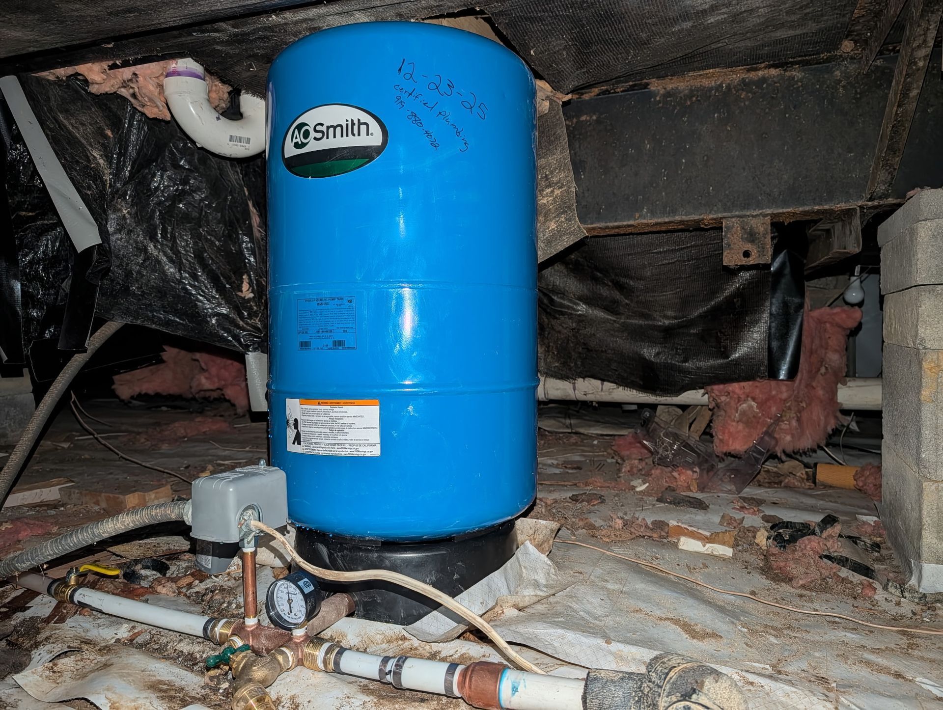 A blue A.O. Smith well water pressure tank situated in a crawl space with exposed insulation, pipes, and a pressure gauge.