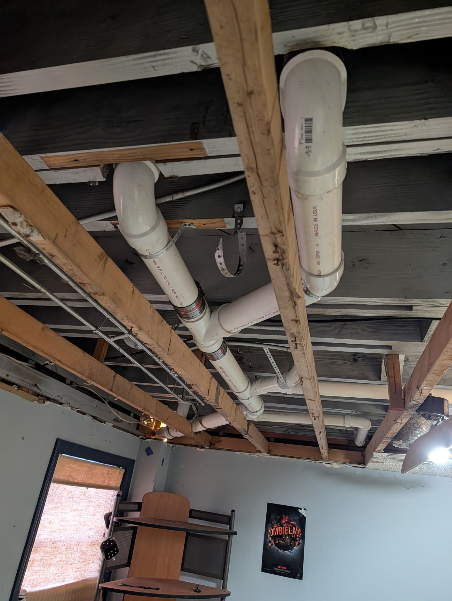 White PVC drainage pipes run across the open-joist ceiling of a room, connected by a Y-shaped fitting.