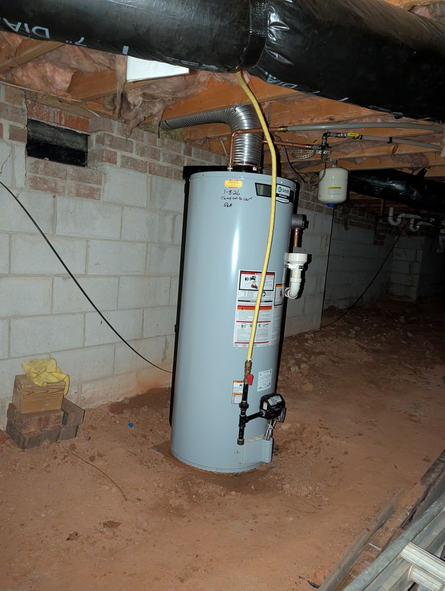 A gray water heater stands on a dirt floor in a basement with exposed brick walls and overhead wooden joists.