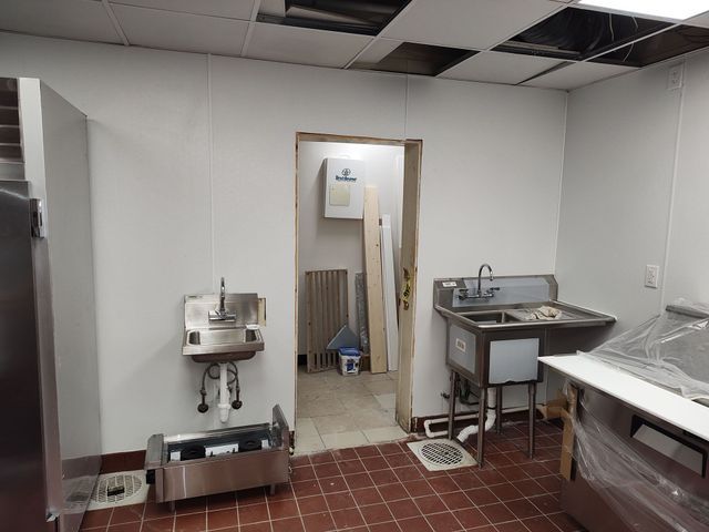 Interior of a commercial kitchen under construction. Walls are white, floor is red tile. Sinks, fixtures, and appliances are visible.