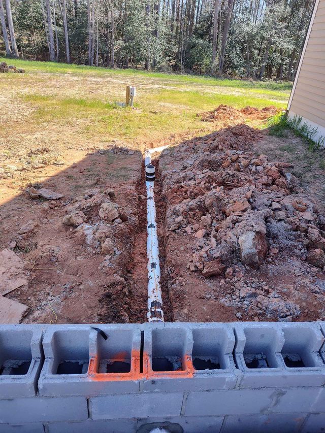Trench with white pipe, alongside a cinder block wall, outdoors. Brown soil, grass, and trees in the background.