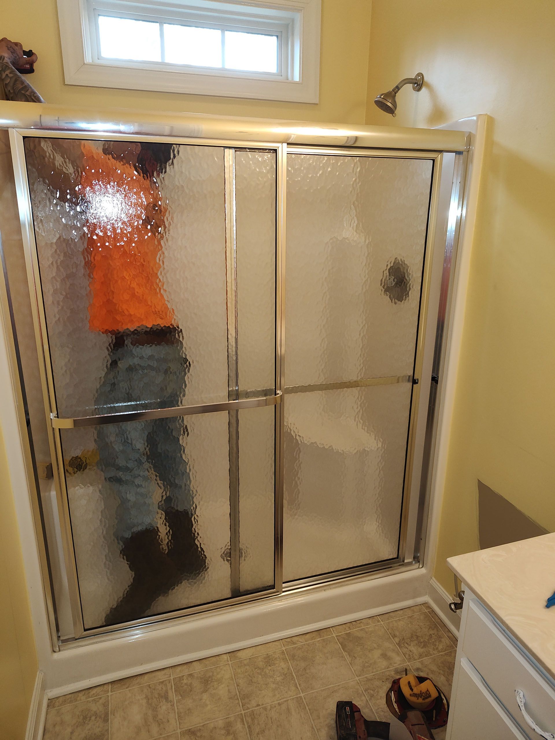 Person inside a shower stall, reaching up. They wear an orange shirt, jeans, and work boots.