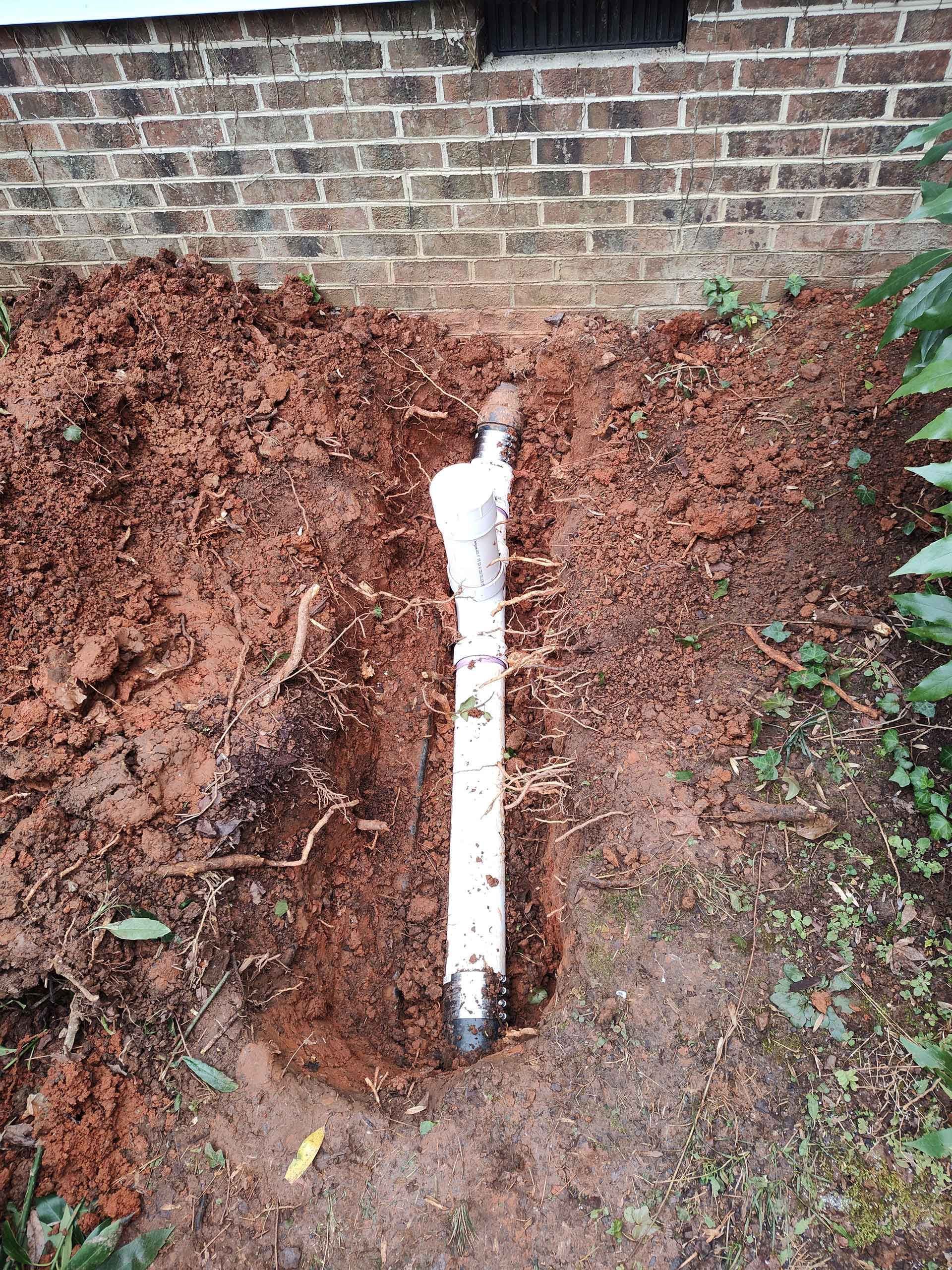 White PVC pipe partially buried in red soil next to a brick wall.