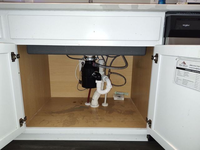 Open white cabinet under a kitchen sink. Garbage disposal and plumbing visible.