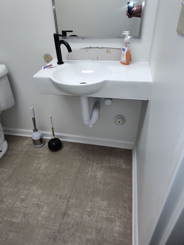 White wall-mounted bathroom sink with black faucet, below a mirror. A toilet is visible on the left.