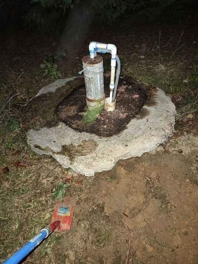 Well pump with concrete base in grassy area. Blue pipe and wiring visible.