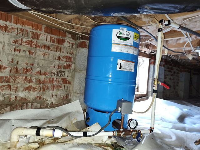 Blue water pressure tank in a crawl space, against a brick wall, with plumbing and pressure gauge.