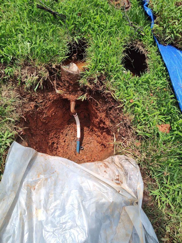 Hole in the ground with exposed pipe and a blue handle, surrounded by grass and dirt.