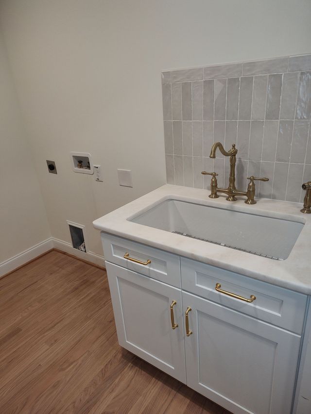 White laundry sink with gold faucet and hardware, white cabinet, light tile backsplash, hardwood floor.