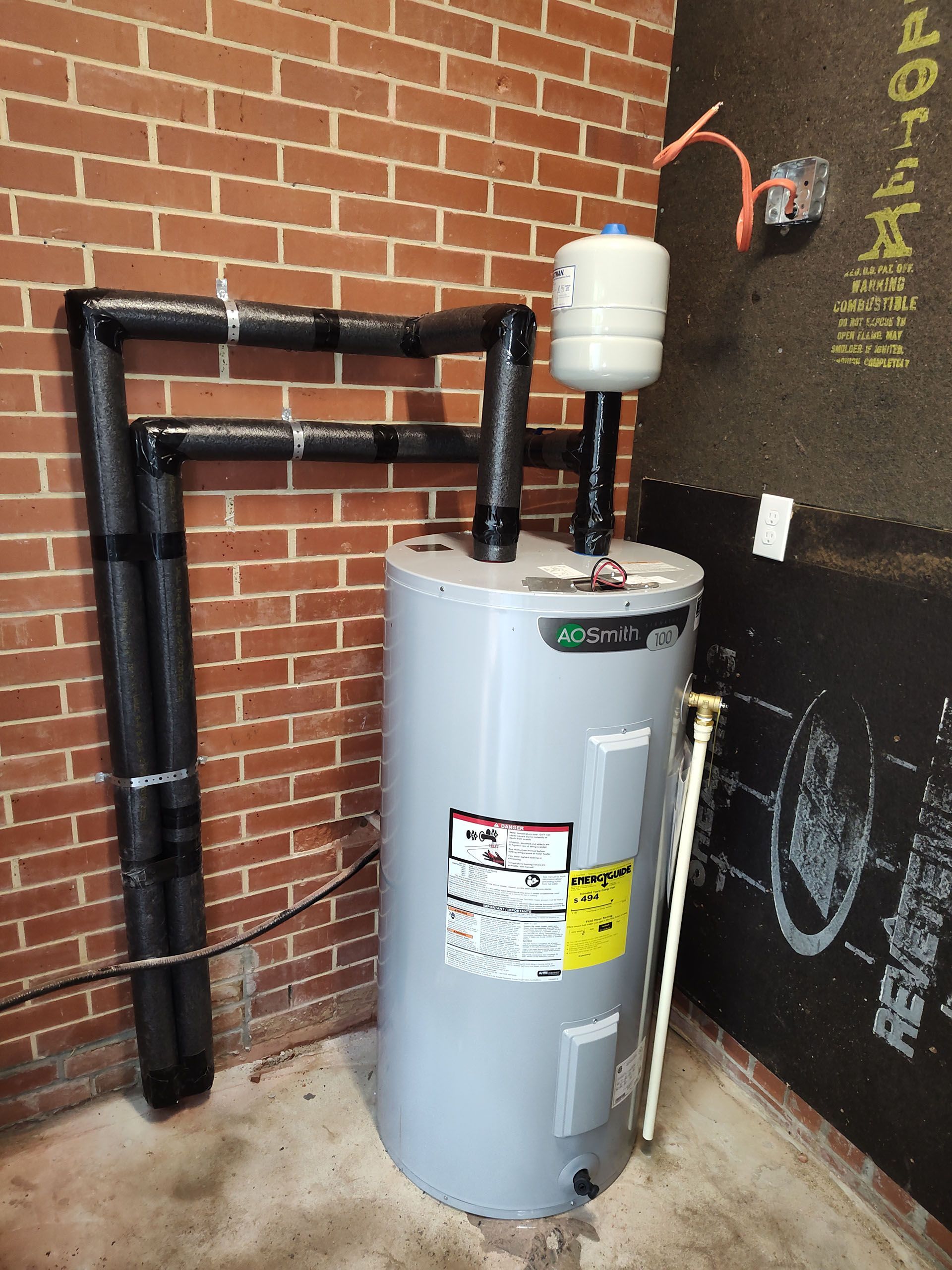 Water heater with black insulated pipes against a brick wall. Expansion tank and electrical components visible.