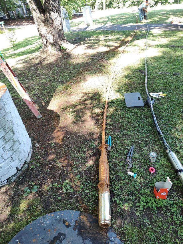 Rusty well pump on the ground near a well head, electrical wiring, and a person in the background.