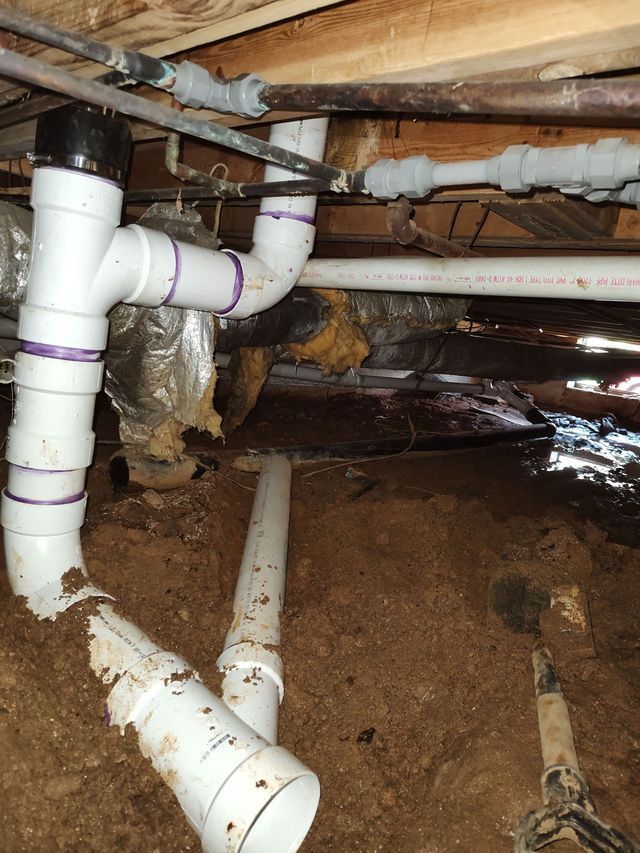 Underneath a structure: white PVC pipes and copper pipes in a crawl space, with dirt and insulation visible.