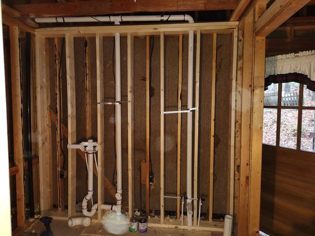 Interior view of a bathroom remodel in progress; wood framing, exposed pipes, and plumbing fixtures.