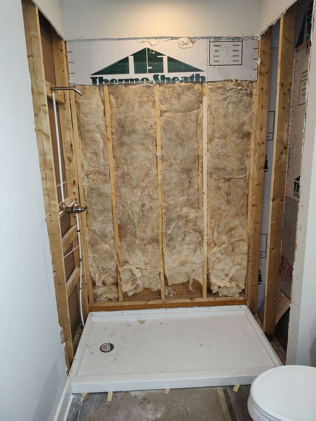 Shower stall under construction, exposed framing with insulation. White shower base and toilet visible.