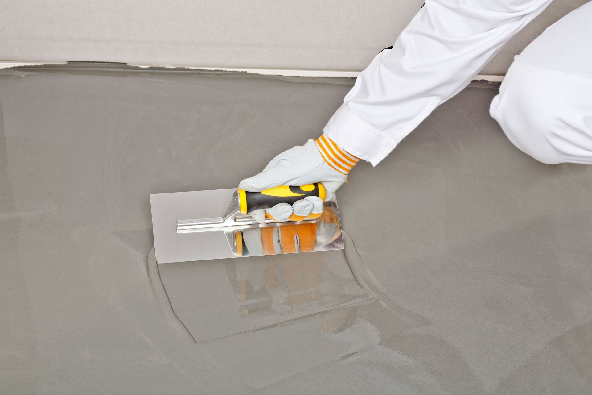 Person smoothing gray cement with a trowel on a floor, wearing gloves and white sleeves.