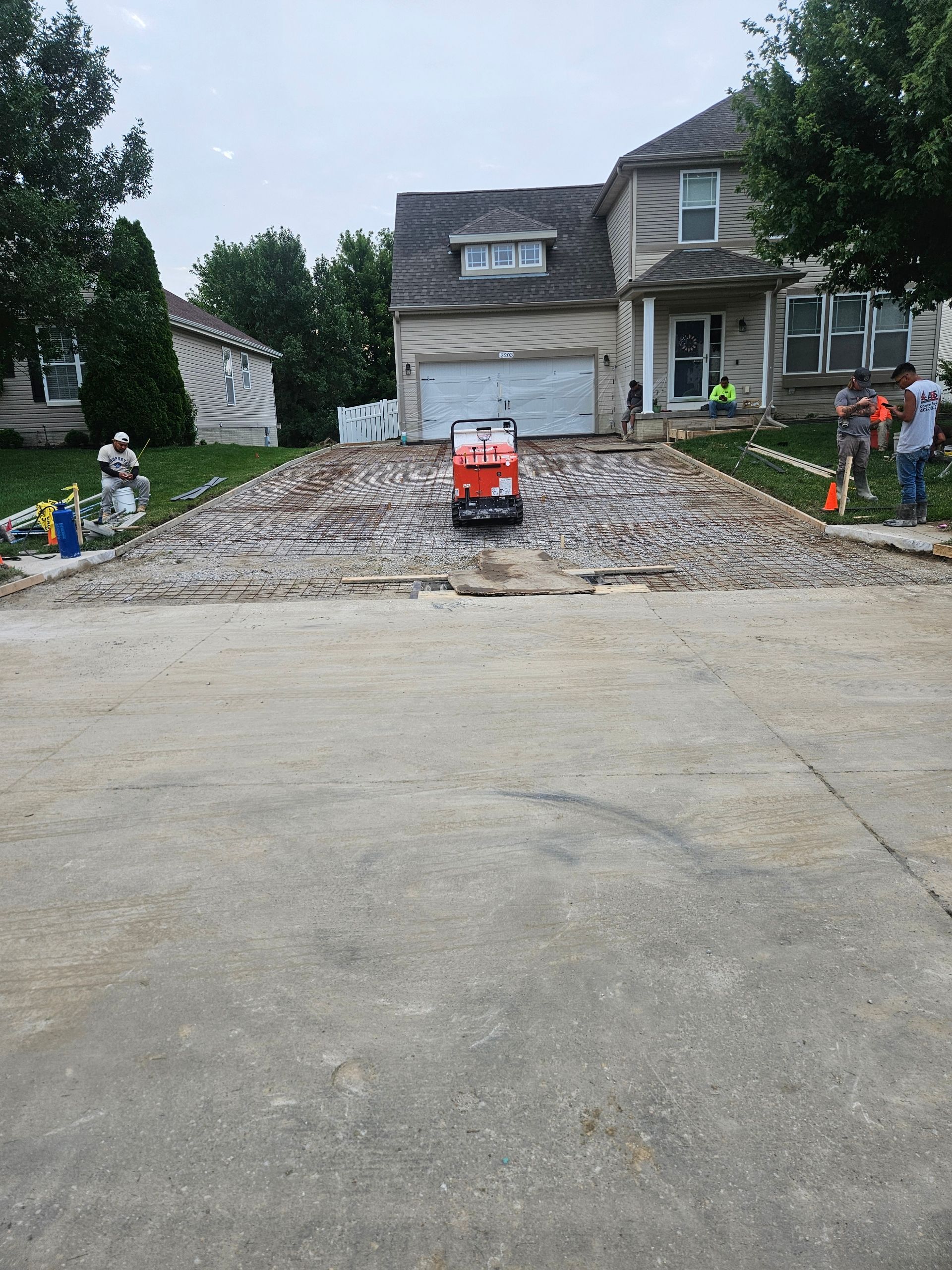 Driveway construction at a two-story house. Red compactor machine on the rough surface, workers nearby.