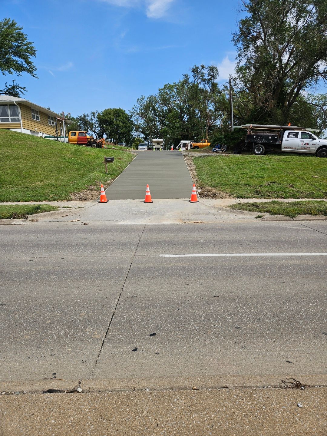 New concrete pathway under construction, bordered by grass, with orange cones on a road.