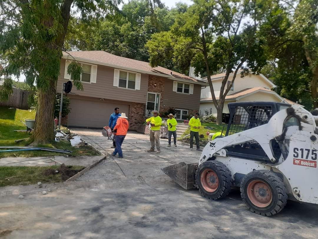 Workers paving a driveway in front of a two-story beige house; Bobcat loader present.