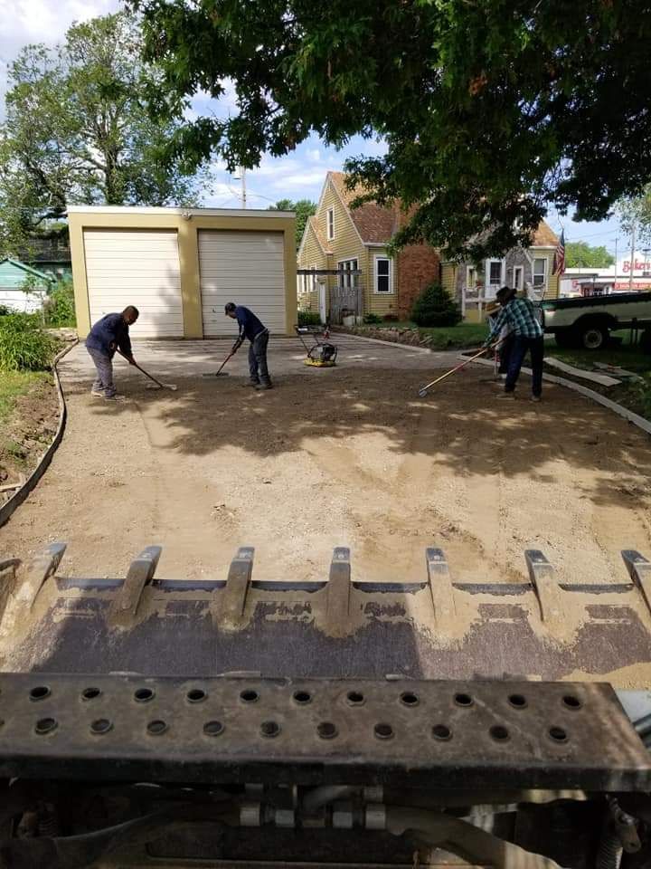 Three people raking gravel on a driveway, with a garage and house in the background.