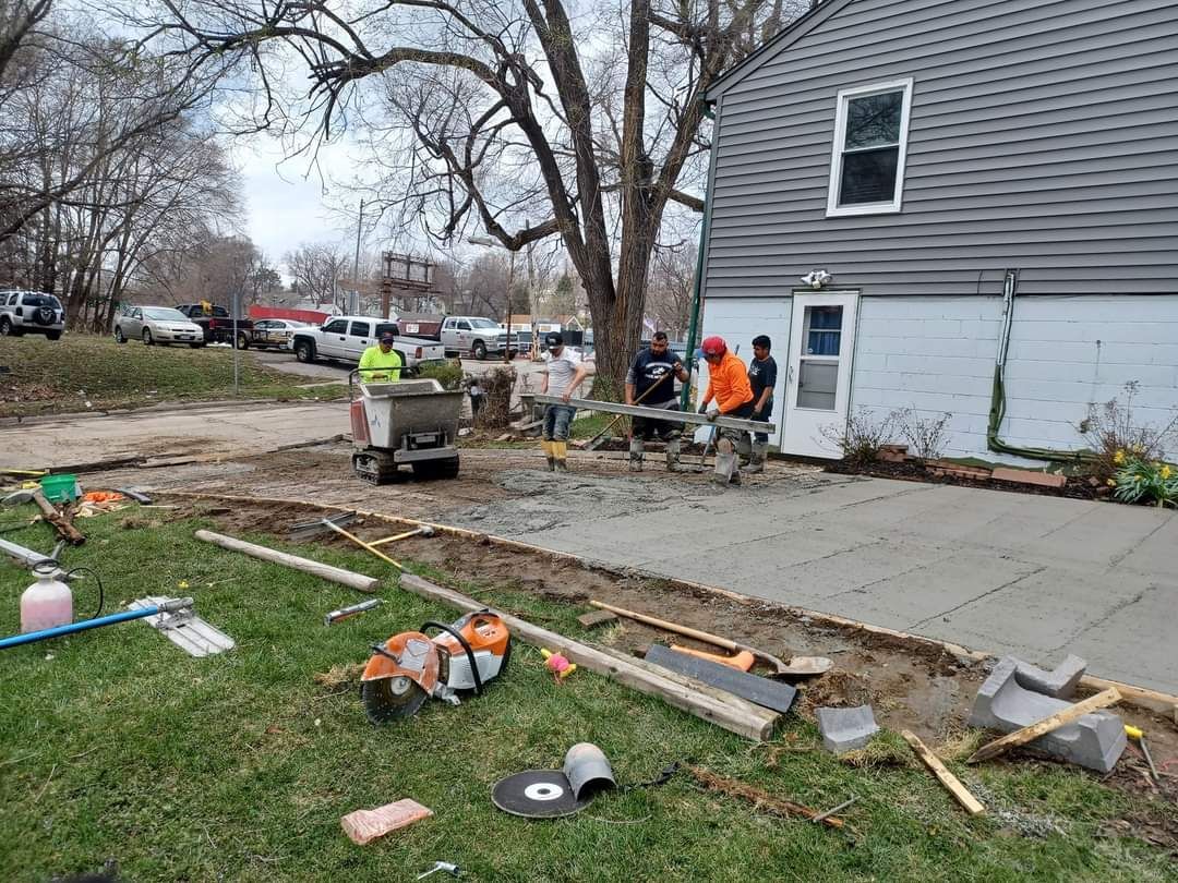 Construction workers pouring concrete on a driveway next to a gray house; various tools and materials are visible.