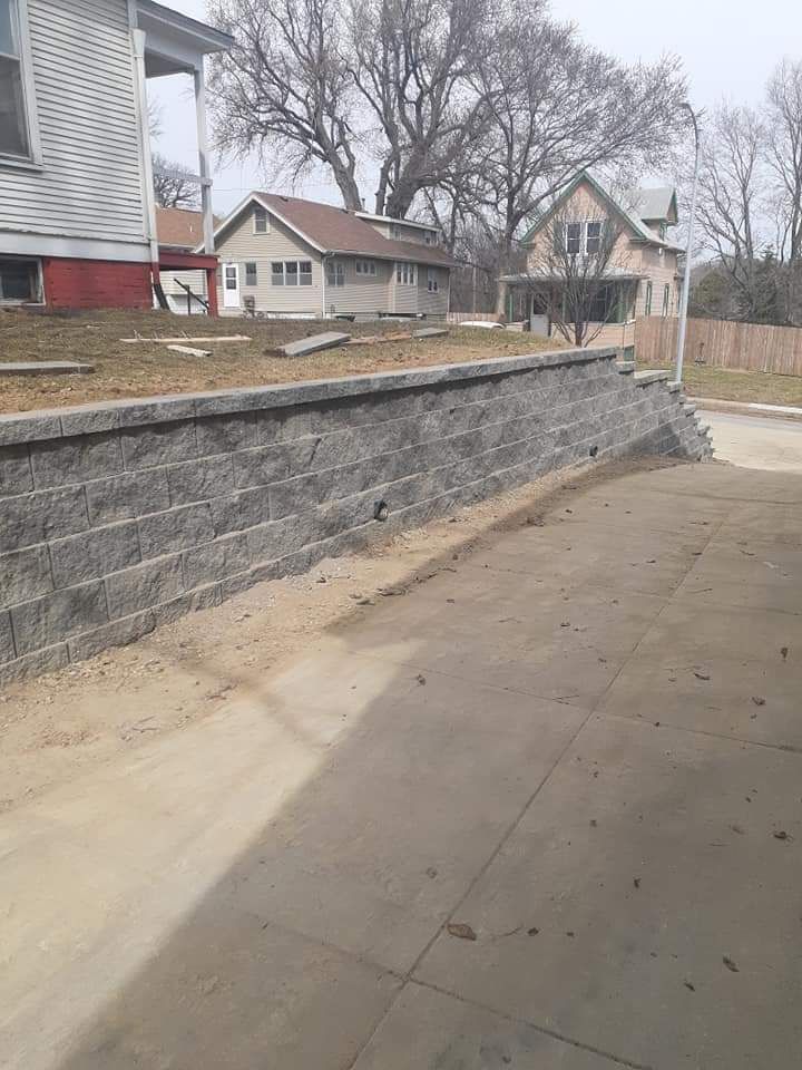 Gray retaining wall next to a concrete driveway, with houses and trees in the background.