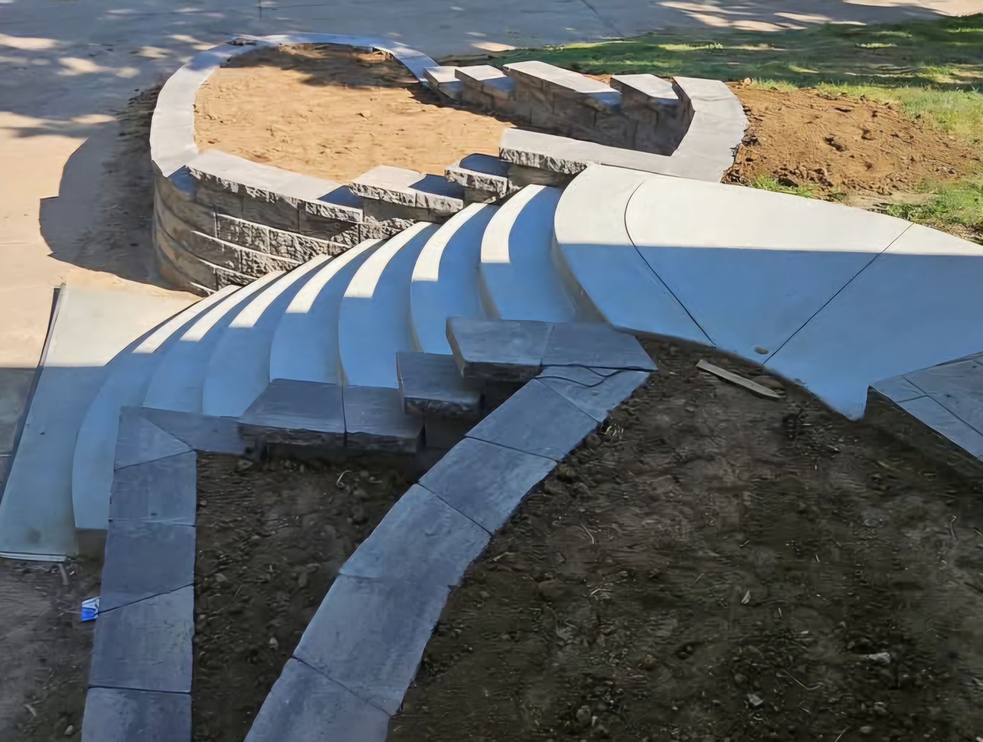 Curved stone steps and retaining wall leading to a circular patio under construction; dirt and lawn surround.