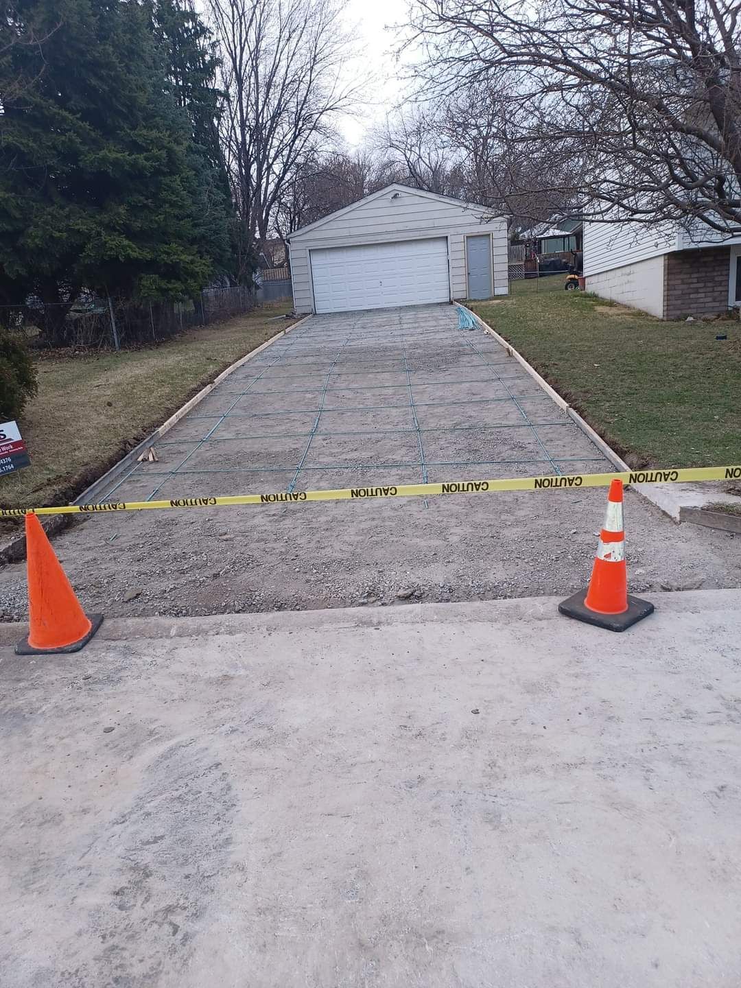 Driveway under construction, gravel surface, orange cones, caution tape, garage in background.